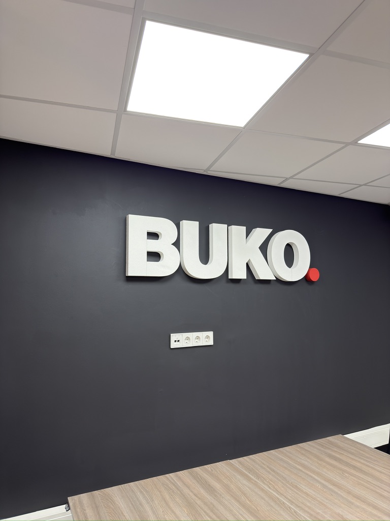 Ruimtelijk wit BUKO-logo met rode punt op een donkere kantoorwand