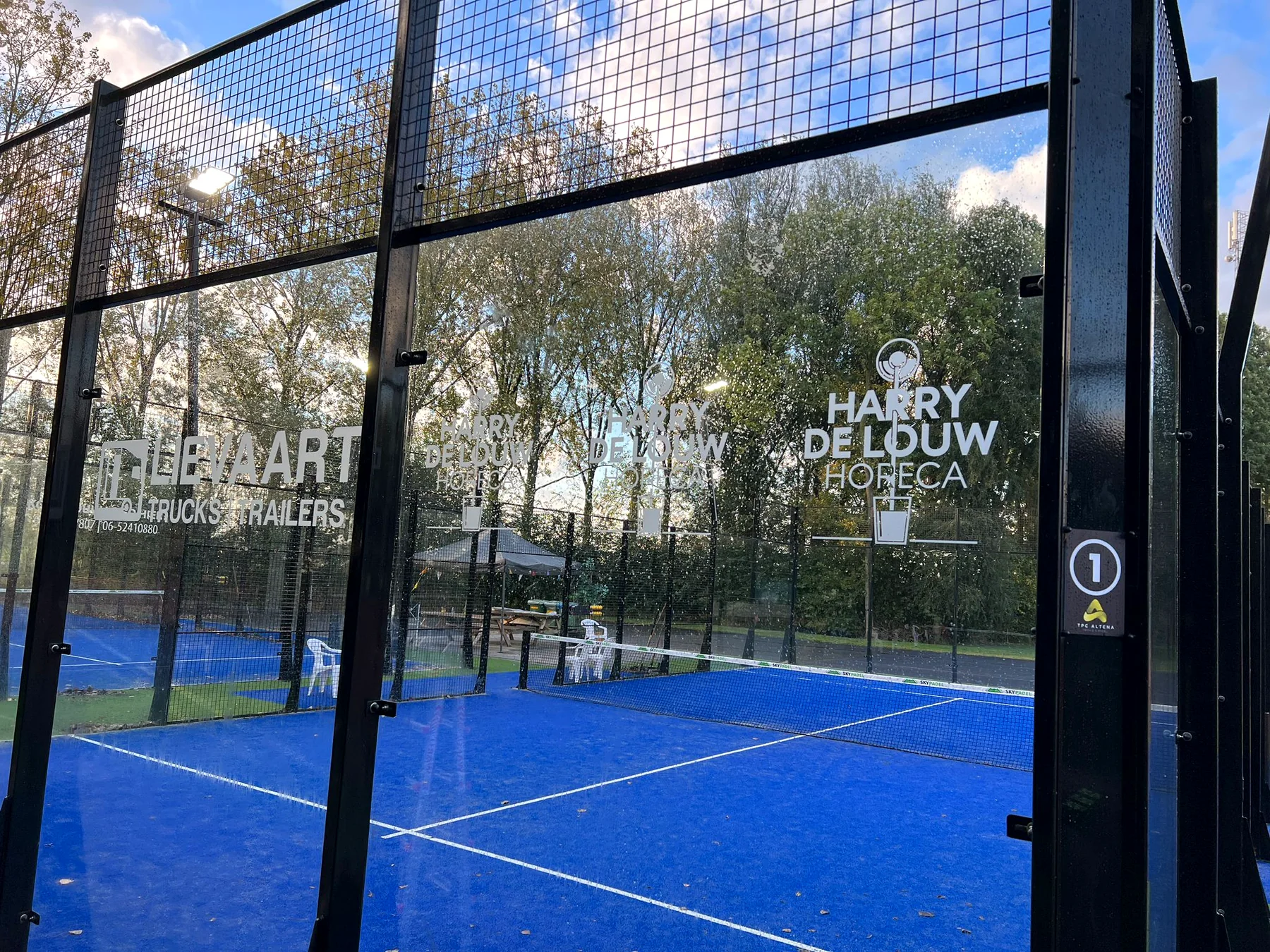 Sponsorstickers van meerdere bedrijven op de glazen wanden van een padelbaan bij TPC Altena