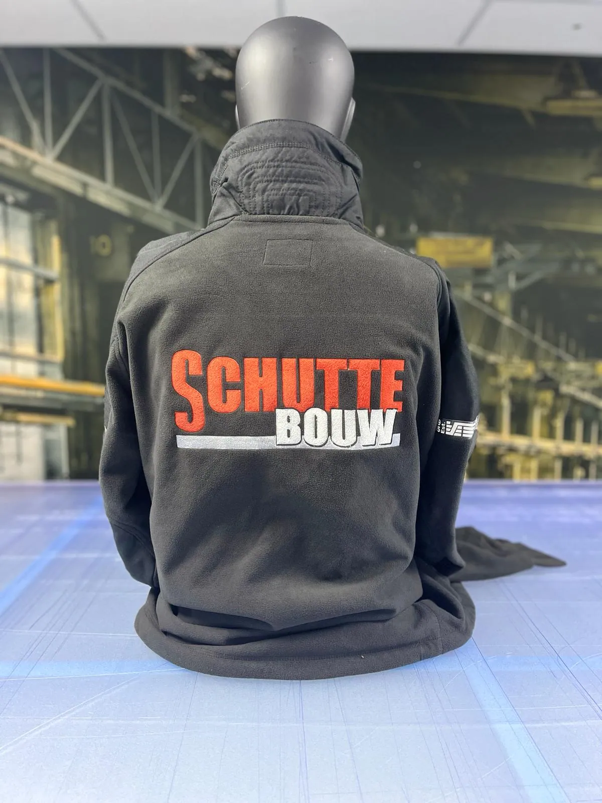 Zwarte werkjas van Schutte Bouw met grote oranje-witte borduring